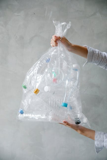 A person's hand is shown holding a semi-transparent green plastic rubbish bag with a knotted top, filled with assorted waste materials. The bag appears full, with various shapes and materials inside,